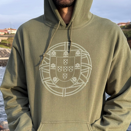 Person wearing a green hoodie with a white portuguese "brasão de armas" emblem, standing by a body of water.