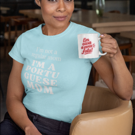 Woman wearing a light blue t-shirt with "I'm nor a regular a regular mom I'm a portuguese mom"text, holding a mug in a casual setting.