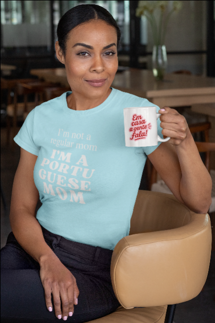 Woman wearing a light blue t-shirt with "I'm nor a regular a regular mom I'm a portuguese mom"text, holding a mug in a casual setting.