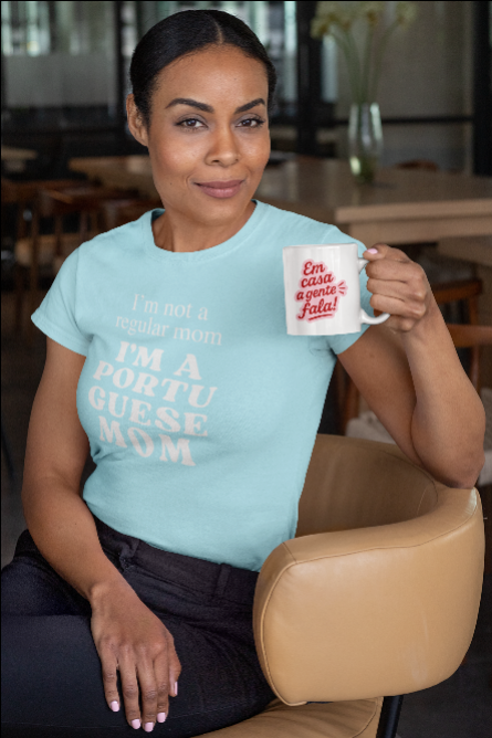 Woman wearing a light blue t-shirt with "I'm nor a regular a regular mom I'm a portuguese mom"text, holding a mug in a casual setting.