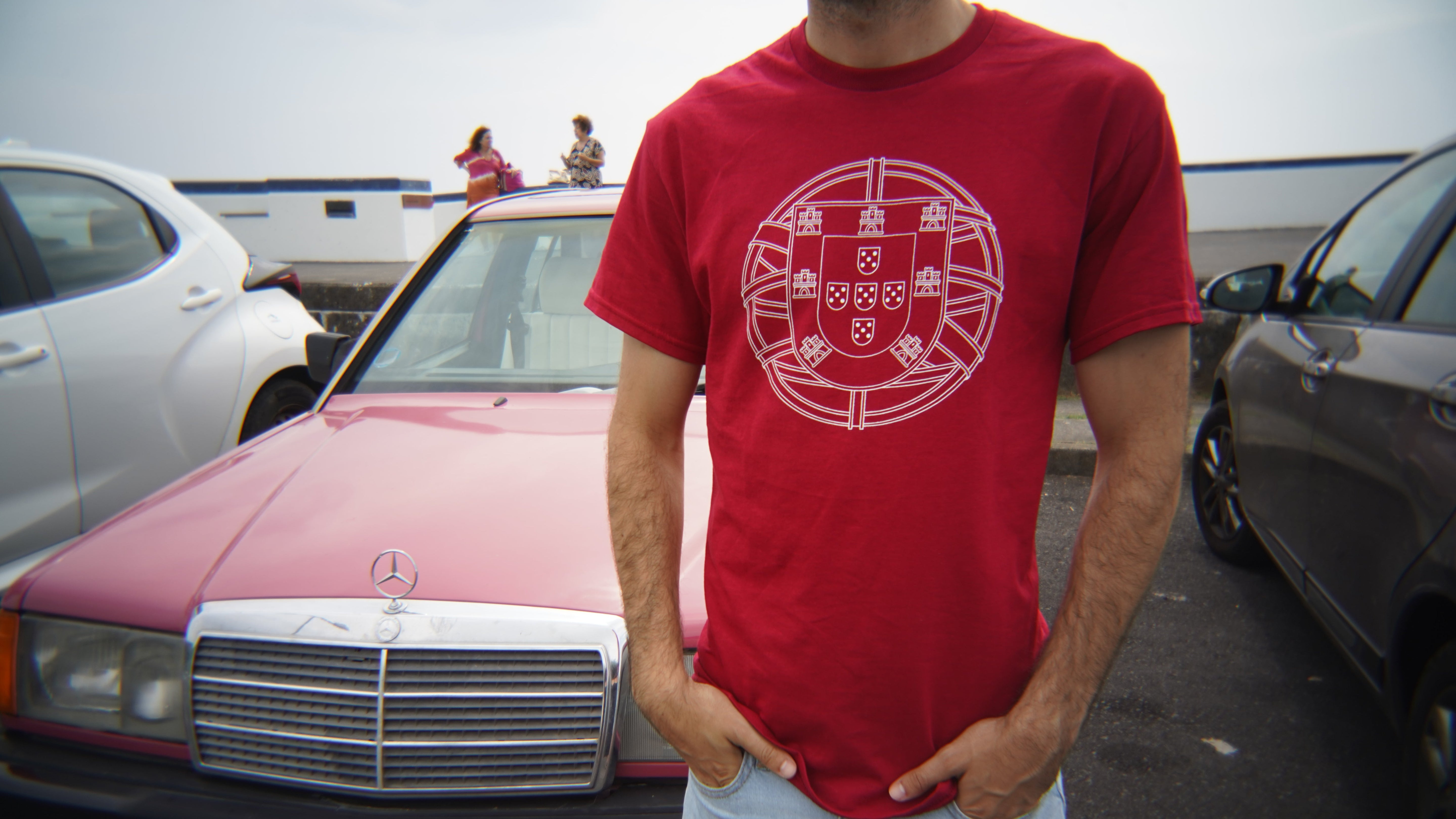 Person wearing a red t-shirt with a white portuguese "brasão de armas" in front of a pink Mercedes-Benz car.