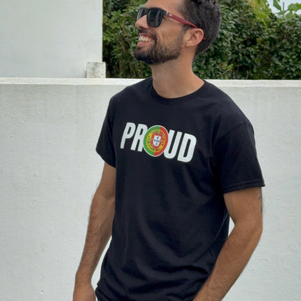 Man wearing a black t-shirt with 'PROUD to be portuguese' text, sunglasses, and light jeans standing against a white wall with greenery.