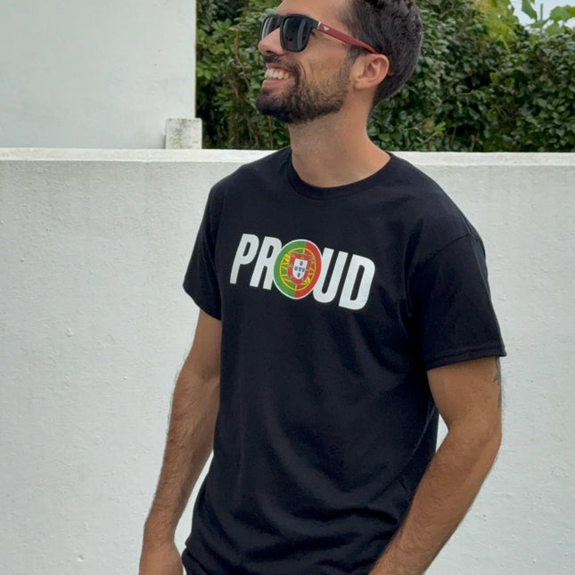 Man wearing a black t-shirt with 'PROUD to be portuguese' text, sunglasses, and light jeans standing against a white wall with greenery.