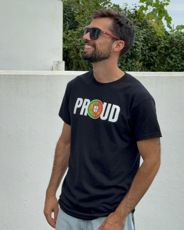 Man wearing a black t-shirt with 'PROUD to be portuguese' text, sunglasses, and light jeans standing against a white wall with greenery.