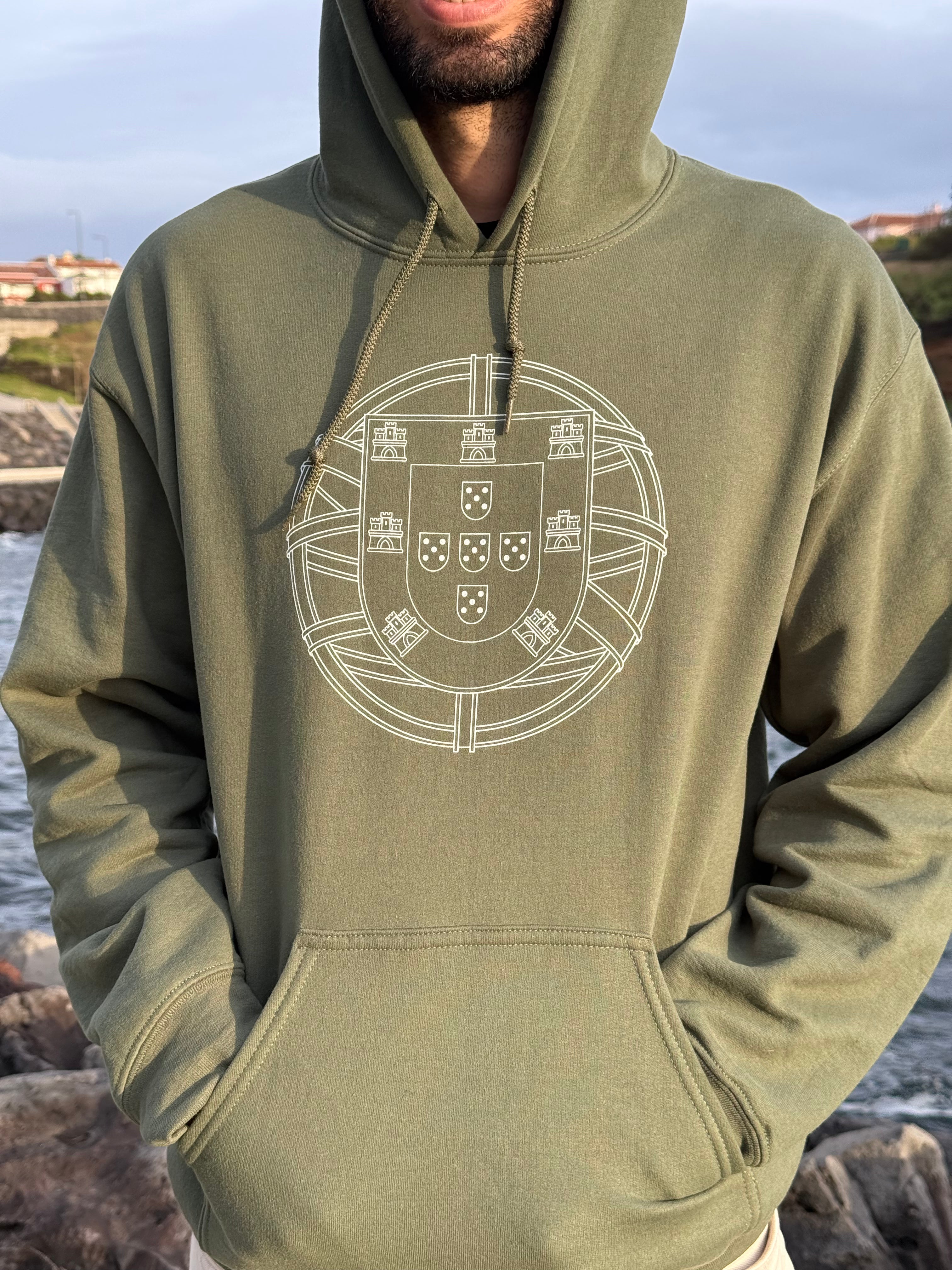 Person wearing a green hoodie with a white portuguese "brasão de armas" emblem, standing by a body of water.
