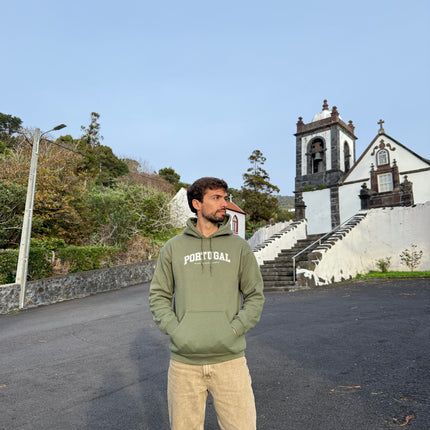 Man wearing a green hoodie with 'Portugal, nação valente e imortal' text standing on a road with a church in the background.