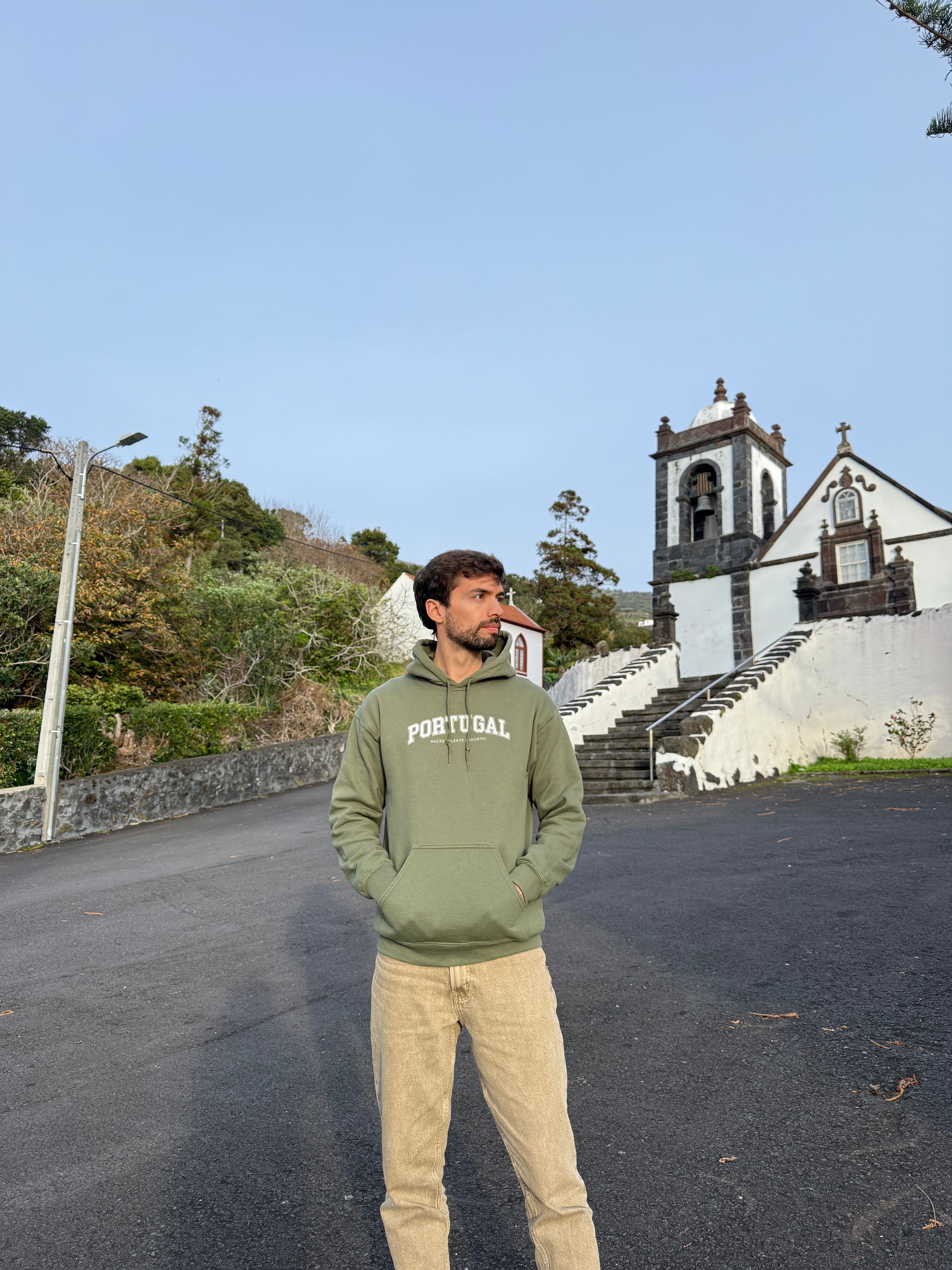Man wearing a green hoodie with 'Portugal, nação valente e imortal' text standing on a road with a church in the background.