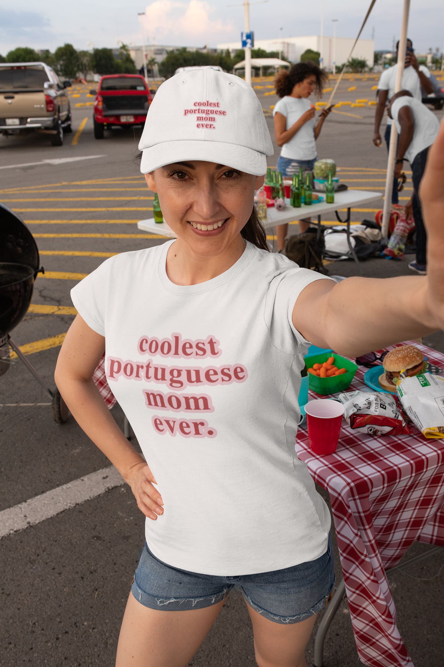 Woman wearing a white t-shirt with 'coolest portuguese mom ever' text, taking a selfie in a parking lot.