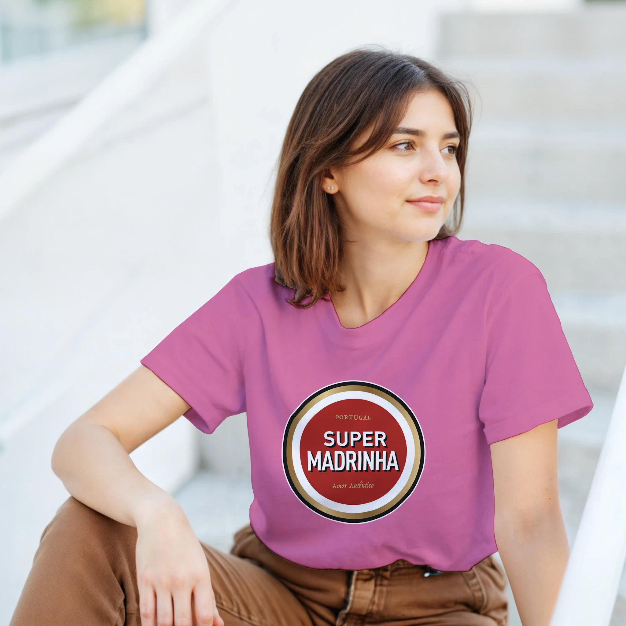 Woman wearing a pink t-shirt with 'Super Madrinha' design sitting outdoors.