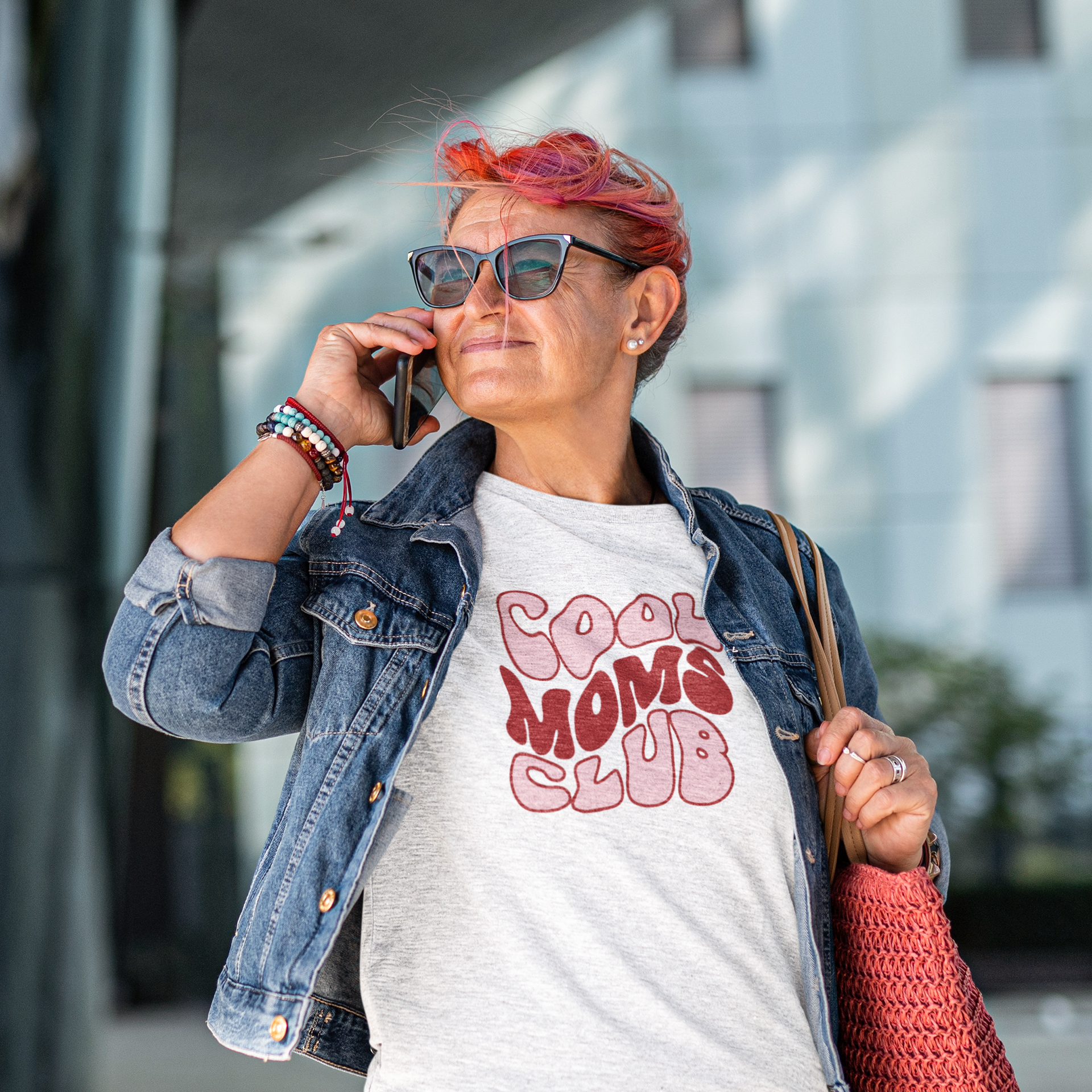 portuguese woman wearing a 'Cool Moms Club' t-shirt, talking on a phone outdoors.